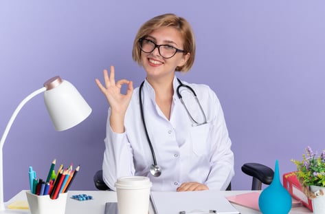 smiling-young-female-doctor-wearing-medical-robe-with-stethoscope-glasses-sits-table-with-medical-tools-showing-okay-gesture-isolated-blue-background